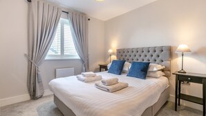 A neatly made bed with white linens and blue pillows, a bedside table with a lamp, and a window with curtains. - 3 Bed in Fowey (oc-s33234) (Fowey)