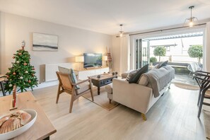 A modern living room with a sofa, coffee table, and a TV. A Christmas tree is visible, and there's a dining area with a table and chairs. - 3 Bed in Fowey (oc-s33234) (Fowey)