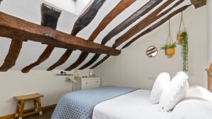 A bedroom with a sloped ceiling, a bed with white bedding, a small wooden stool, a nightstand with a lamp, and a hanging plant. - Casa Rural Selmo in the Heart of Asturias (Zancornio)