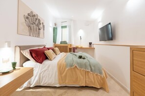 Un dormitorio con cama, mesita de noche, televisor, obra de arte y una ventana con cortinas.