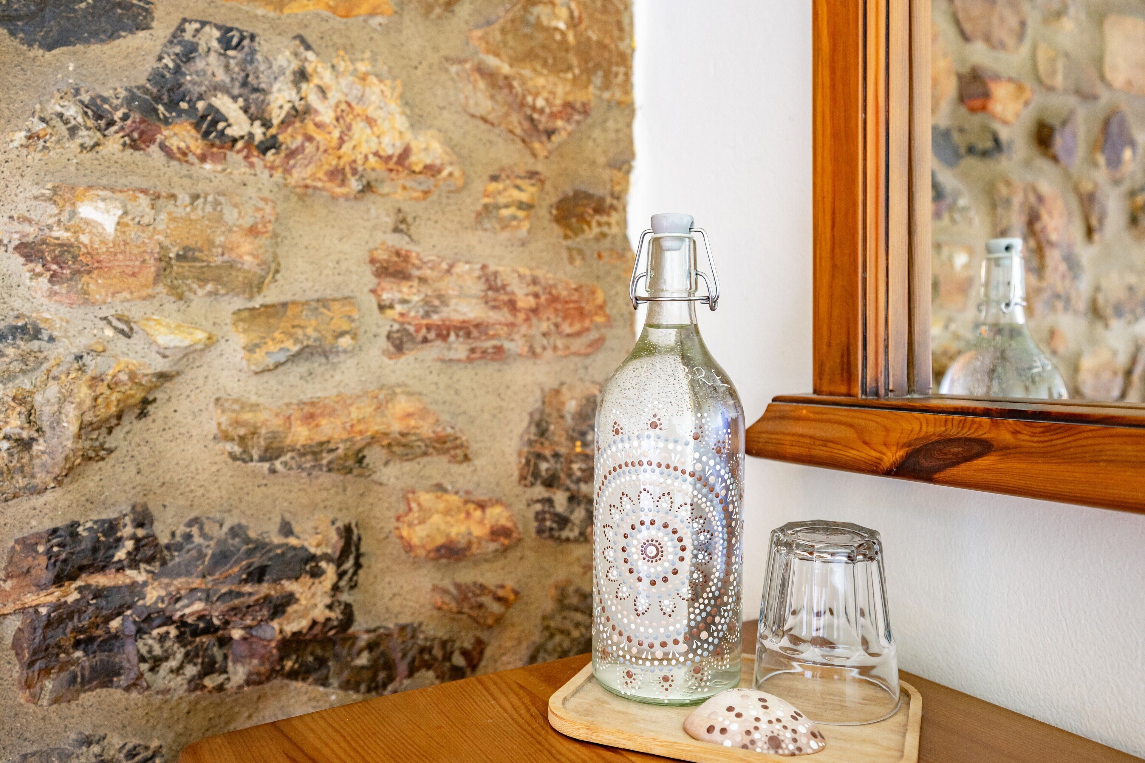 Een fles met een decoratief patroon, een helder glas en een onderzetter op een houten tafel voor een bakstenen muur.