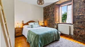A bedroom with a stone wall, wooden floor, a bed with a green bedspread, a wooden nightstand, a lamp, and a window with curtains. - Casa Rural Selmo in the Heart of Asturias (Zancornio)
