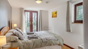 Une chambre à coucher avec un lit, des tables de chevet, une fenêtre avec des rideaux, un tableau au mur et une vue sur les arbres à l’extérieur.