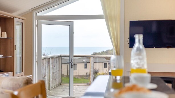 A room with a large sliding glass door leading to a balcony with a view of the sea. There is a flat-screen TV mounted on the wall and a table with a bottle and glasses on it. - 2 Bed in Tresaith (oc-c33408) (Tresaith)