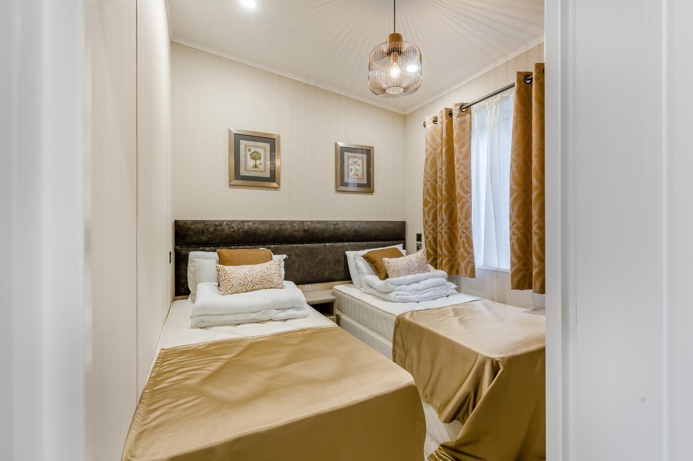 A hotel room with two beds, a large window with curtains, a ceiling light, and two framed pictures on the wall.