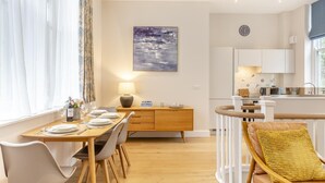 A modern kitchen with white cabinets, a dining area with a wooden table and chairs, and a yellow cushion on a wicker chair. - 2 Bed in Fowey (oc-n33435) (Fowey)
