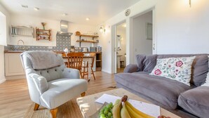 Una sala de estar con un sofá gris, un sillón de madera y una mesa de centro con fruta. Se ve una cocina al fondo, con un comedor.