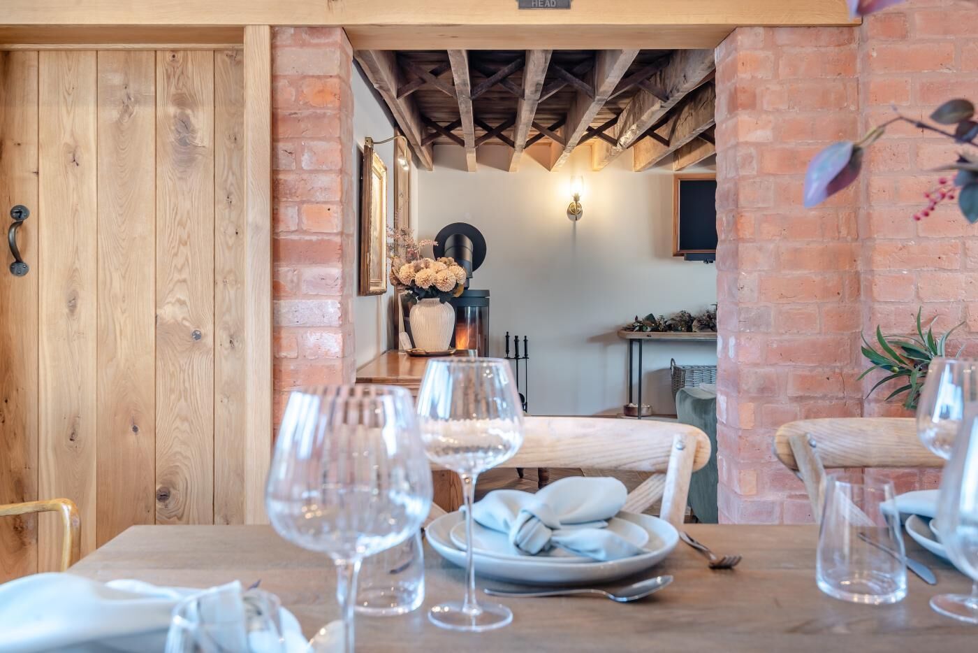A dining area with a brick wall, wooden ceiling beams, and a table set for two with wine glasses and plates.