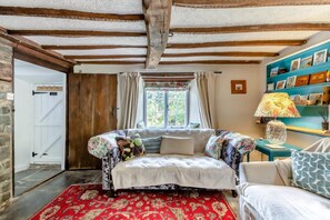 Een gezellige kamer met zichtbare houten balken, een bed met bloemenbedekking, een rood gestreept vloerkleed en een blauwgroene bijzettafel met een lamp.