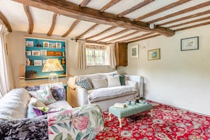 Een gezellige kamer met een rood, gestreept tapijt, een witte zithoek, een blauwgroene plank met diverse spullen, en een houten plafond.