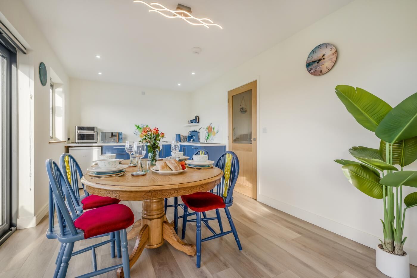 A dining area with a round table, blue chairs, and red cushions. There is a kitchen in the background with modern appliances.