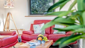 Een moderne woonkamer met een rode bank, een houten salontafel met fruit en tijdschriften, een vloerlamp en uitzicht op bomen door een raam.
