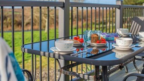 Een glazen tafel gedekt met theekoppen, borden en een brood op een terras met een metalen hekwerk.