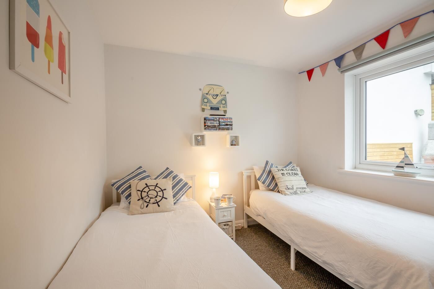 A bedroom with two single beds, a small bedside table with a lamp, a shelf with books, and a window with a view of the outside.