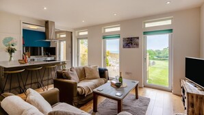 A modern living room with a sofa, coffee table, and a TV. There is a kitchen area with a bar and a dining table. Large windows provide a view of the outdoors. - 2 Bed in Harrowbarrow (oc-w33960) (Harrowbarrow)