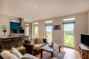 Ein modernes Wohnzimmer mit einer Couch, einem Couchtisch und einem Fernseher. Es gibt eine Küchenzeile mit einer Bar und einem Esstisch. Große Fenster bieten einen Blick ins Freie.