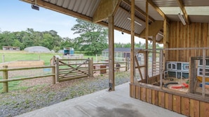 Ein überdachter Außenbereich mit Holzkonstruktion, einem verschlossenen Eingang und Blick auf eine Farm mit grünen Feldern und Bäumen.