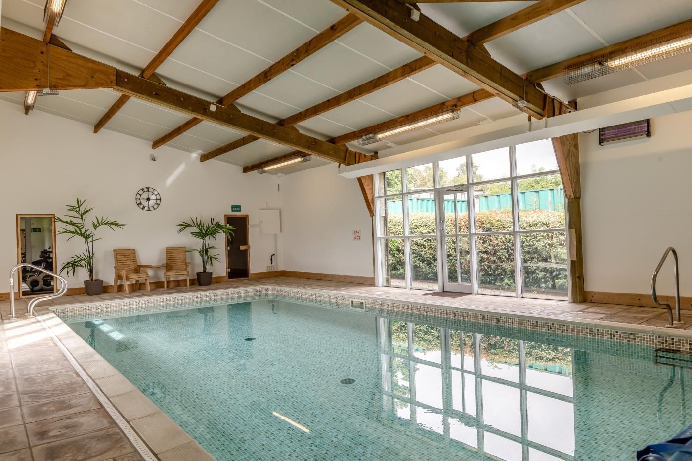 An indoor swimming pool with wooden beams, a clock, and potted plants.