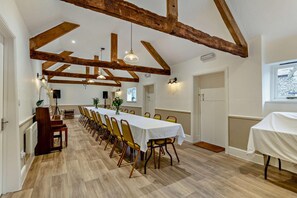 Une longue table à manger entourée de chaises, des poutres en bois et des luminaires suspendus dans une pièce spacieuse dotée d’un plancher en bois.