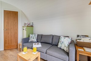 Un salon chaleureux avec un canapé gris, des coussins à motifs floraux, une table basse en bois sur laquelle reposent deux verres de jus d’orange, et un panier en osier posé sur une table d’appoint en bois.