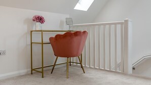 Ein Zimmer mit weißer Decke, einem Oberlicht, einer weißen Brüstung, einem goldfarbenen Schreibtisch, einem rosafarbenen Sessel und einer Vase mit Blumen.
