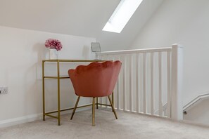 Een kamer met een wit plafond, een dakraam, een witte balustrade, een bureau met gouden lijst, een roze stoel en een vaas met bloemen.