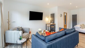 Ein modernes Wohnzimmer mit einem grauen Sofa, einem blauen Sofa, einem kleinen runden Tisch mit einer Lampe und einem an der Wand befestigten Fernseher.