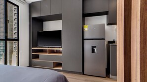 Quarto moderno com cama, TV integrada e pequena cozinha compacta.