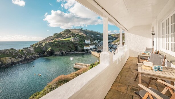 A coastal view from a balcony with a table and chairs, overlooking a bay with a small boat and houses on a hillside. - 3 Bed in Polperro (oc-h34100) (Polperro)