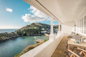 A coastal view from a balcony with a table and chairs, overlooking a bay with a small boat and houses on a hillside. - 3 Bed in Polperro (oc-h34100) (Polperro)