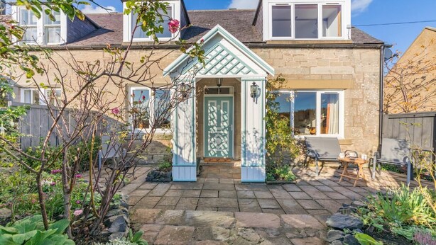 Ein zweistöckiges Haus mit einer Steinaußenwand, einer Veranda mit einer grünen Tür und einem Garten mit verschiedenen Pflanzen und einer Bank.