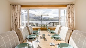 A dining area with a table set for four, large windows with curtains, and a view of a body of water and mountains. - 3 Bed in Lochearnhead (oc-c33641) (Lochearnhead)