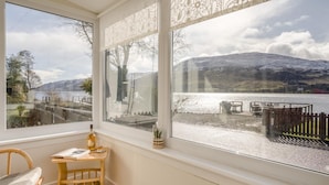 A room with a large window overlooking a lakeside view, a wooden chair, and a small table with a bottle and glass. - 3 Bed in Lochearnhead (oc-c33641) (Lochearnhead)
