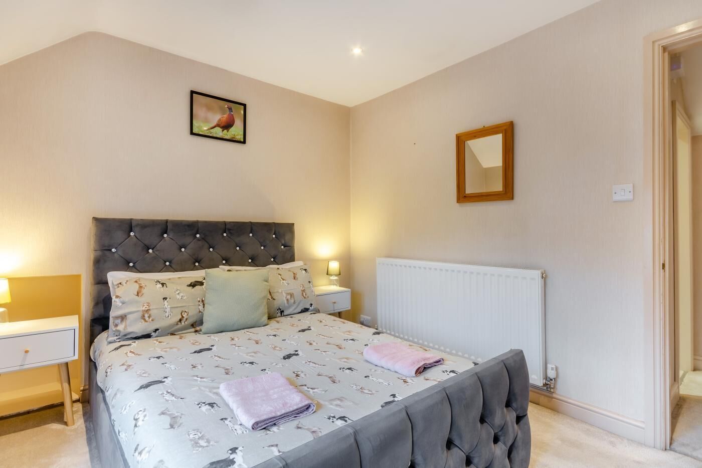 Une chambre à coucher avec une tête de lit capitonnée, un lit recouvert d’une literie à motifs floraux, une table de chevet avec une lampe, un radiateur et un tableau encadré accroché au mur.