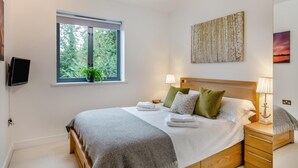 Une chambre avec une grande fenêtre, un lit avec des oreillers et une couverture, une table de chevet avec une lampe, et un téléviseur fixé au mur.
