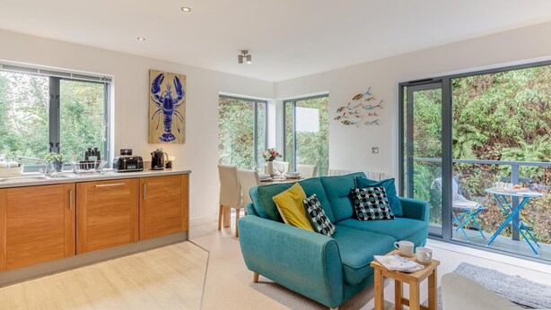 Une cuisine moderne avec des meubles en bois, un coin repas agrémenté d’un canapé turquoise et de grandes fenêtres offrant une vue sur la verdure.