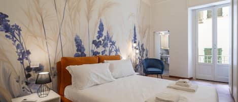 A bedroom with a large bed, a nightstand with a lamp, a chair, and a bathroom visible through an open door.