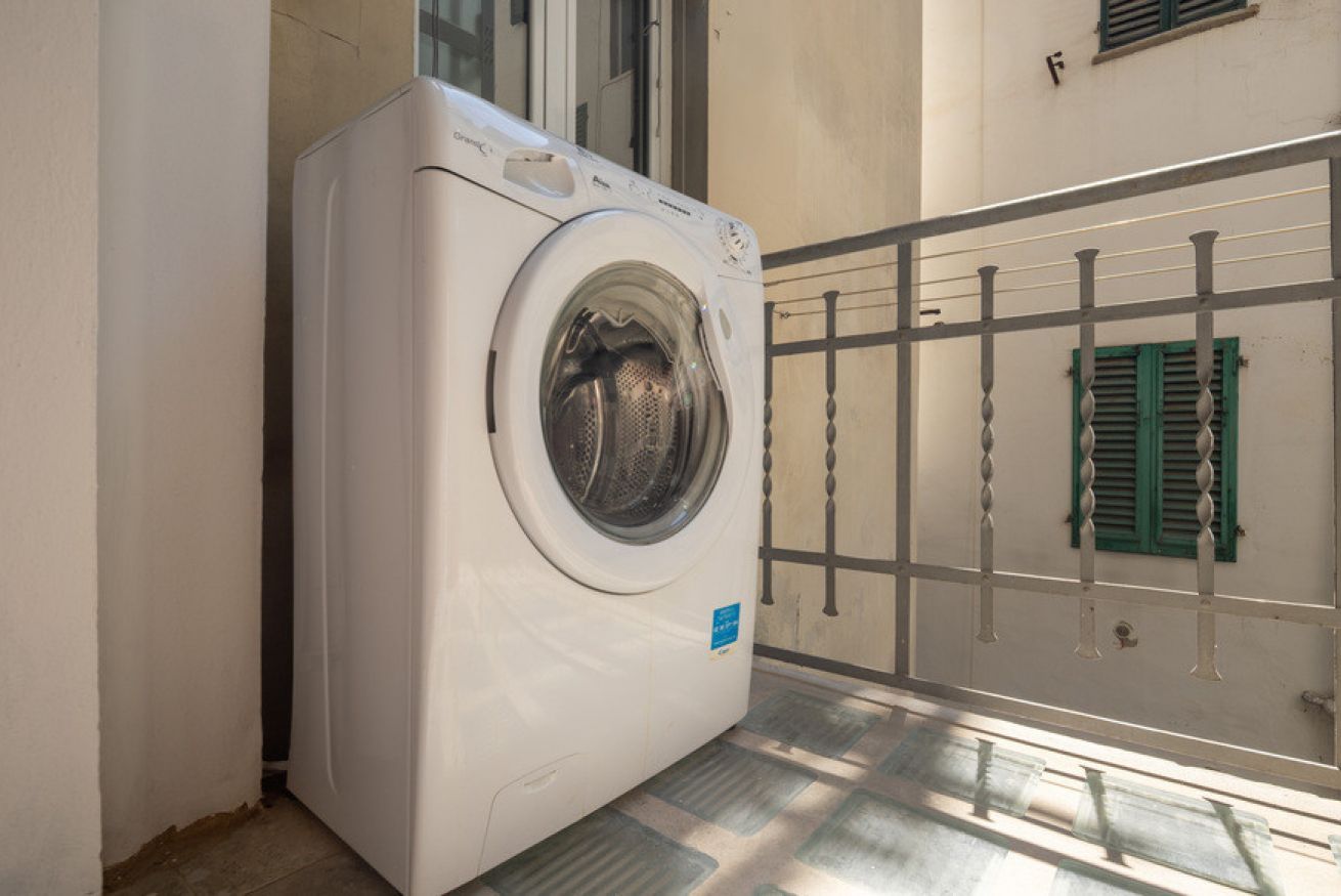 Una lavadora de carga frontal en un cuarto de lavado con una barandilla metálica y una ventana con contraventanas verdes.