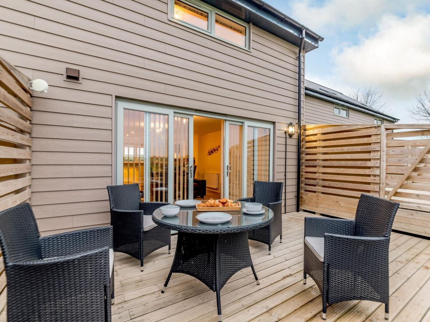 A patio with a round table and four chairs, a sliding glass door leading to an interior room, and a wooden deck.