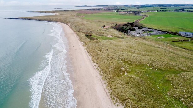 Une vue aérienne d’une zone côtière avec une plage de sable, des champs verdoyants et un ensemble de bâtiments.