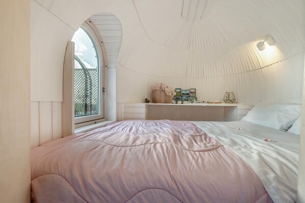 A bedroom with a large window, a bed with a pink bedspread, a bedside table with a lamp, and a wall-mounted shelf with decorative items.