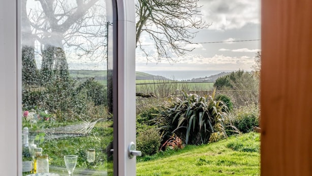 Une table à manger dressée avec des assiettes, des verres et des couverts, un verre d’eau et une bouteille, avec vue sur un paysage verdoyant et des arbres, visible à travers une porte vitrée.