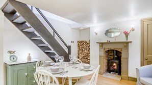Un comedor con una mesa redonda preparada para cuatro personas, sillas blancas, un armario verde, una pared de troncos de madera, una chimenea con un espejo arriba y una escalera a la izquierda.