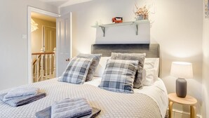 Un dormitorio con cama, almohadas, una lámpara de noche, una estantería con objetos decorativos y una puerta que da a una escalera.