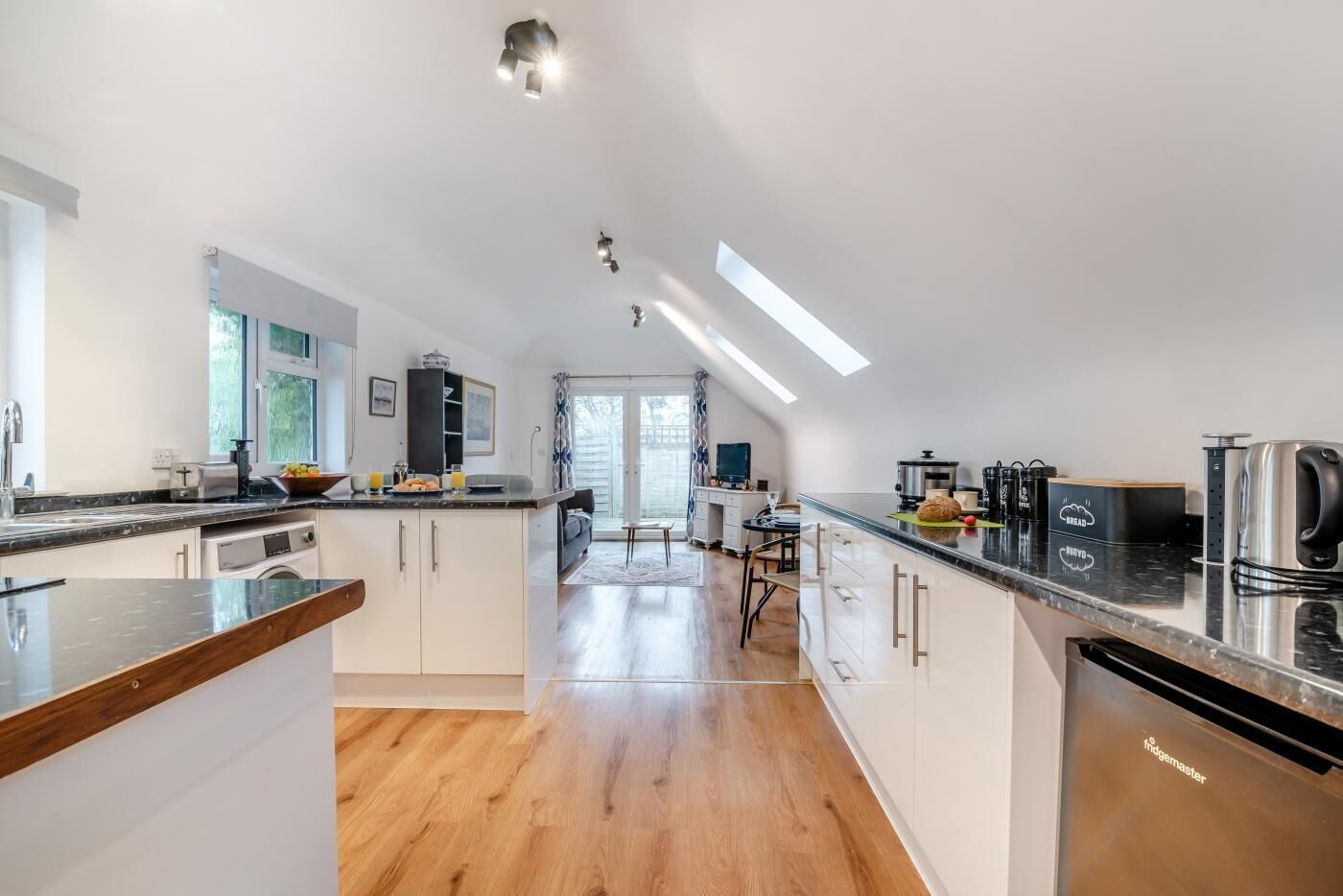 Een moderne keuken met witte kasten, een zwart aanrecht en een roestvrijstalen waterkoker. Er is een eethoek met een tafel en stoelen, en uitzicht op de buitenruimte door een schuifdeur.