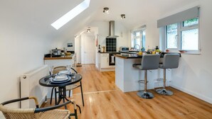 A modern kitchen with a central island, a dining area with a round table, and a view of the adjacent living space. - 1 Bed in Hindhead (oc-w34256) (Haslemere)
