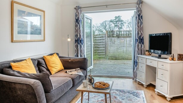 A cozy living room with a grey sofa, yellow cushions, a coffee table with croissants, a TV, and a white dresser. - 1 Bed in Hindhead (oc-w34256) (Haslemere)