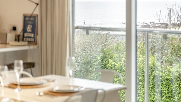 A dining area with a table set for two, a view of the sea, and a sign on the wall. - 4 Bed in Widemouth Bay (oc-s34119) (Widemouth Bay)
