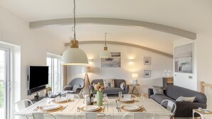 A modern dining area with a white table set for six, surrounded by white chairs. There is a television on the left, a sofa and armchairs in the background, and a large pendant light above the table. - 4 Bed in Widemouth Bay (oc-s34119) (Widemouth Bay)