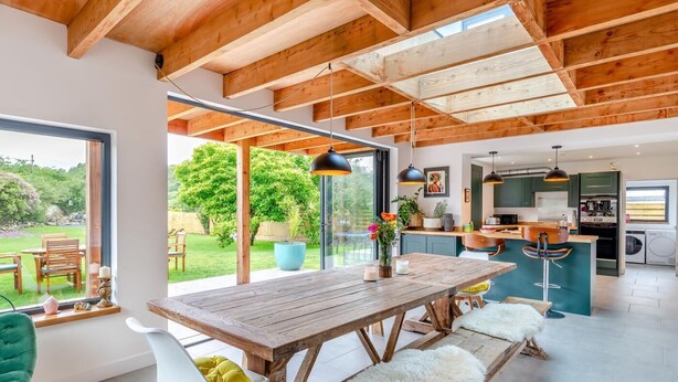 Une cuisine moderne avec des poutres en bois, un coin repas doté d’une grande table et une vue sur un jardin.